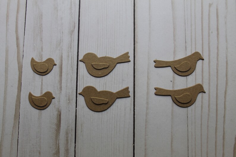 May include: Four brown paper bird cutouts on a white wood background. The birds are different sizes and are arranged in two rows of two.