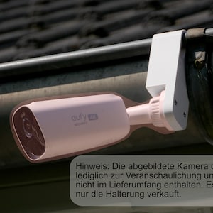 Könnte beinhalten: Eine rosa Sicherheitskamera mit der Aufschrift "eufy 4K SECURITY" ist an einer weißen Halterung befestigt, die an einer dunklen Dachrinne angebracht ist. Die Kamera ist zylindrisch mit einem Objektiv. Der Text im Bild lautet: "Hinweis: Die Kamera dient nur zur Veranschaulichung."