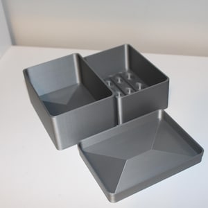 以下が含まれることがあります： グレーの幾何学的な収納ボックスセット。セットには、2つのコンパートメントを備えた長方形のボックス、スロット付きの内部を備えた小さなボックス、および蓋が含まれます。ボックスはマットな素材で作られています。