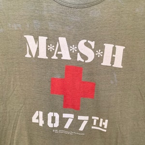May include: Olive green t-shirt featuring the text "M*A*S*H" in white, with asterisks, and a red cross. Below the cross is the number "4077th" in white. The shirt has a crew neck and a vintage look.