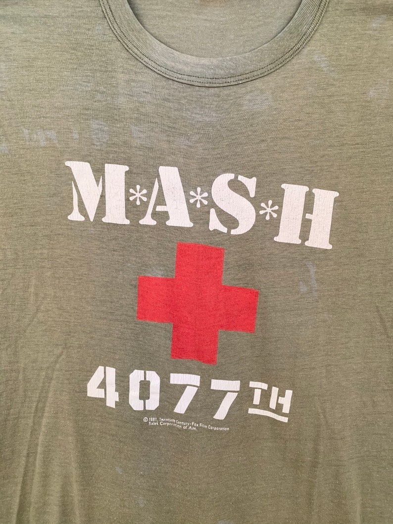 May include: Olive green t-shirt featuring the text "M*A*S*H" in white, with asterisks, and a red cross. Below the cross is the number "4077th" in white. The shirt has a crew neck and a vintage look.