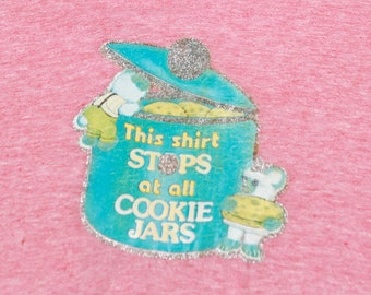 May include: A pink t-shirt with a blue and white cookie jar graphic. The jar has a silver glitter trim and the text "This shirt stops at all cookie jars".