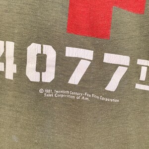 May include: Olive green t-shirt with a large red cross and the white text "4077". Below the text is the copyright information: "&copy; 1981, Twentieth Century-Fox Film Corporation Sales Corporation of Am."