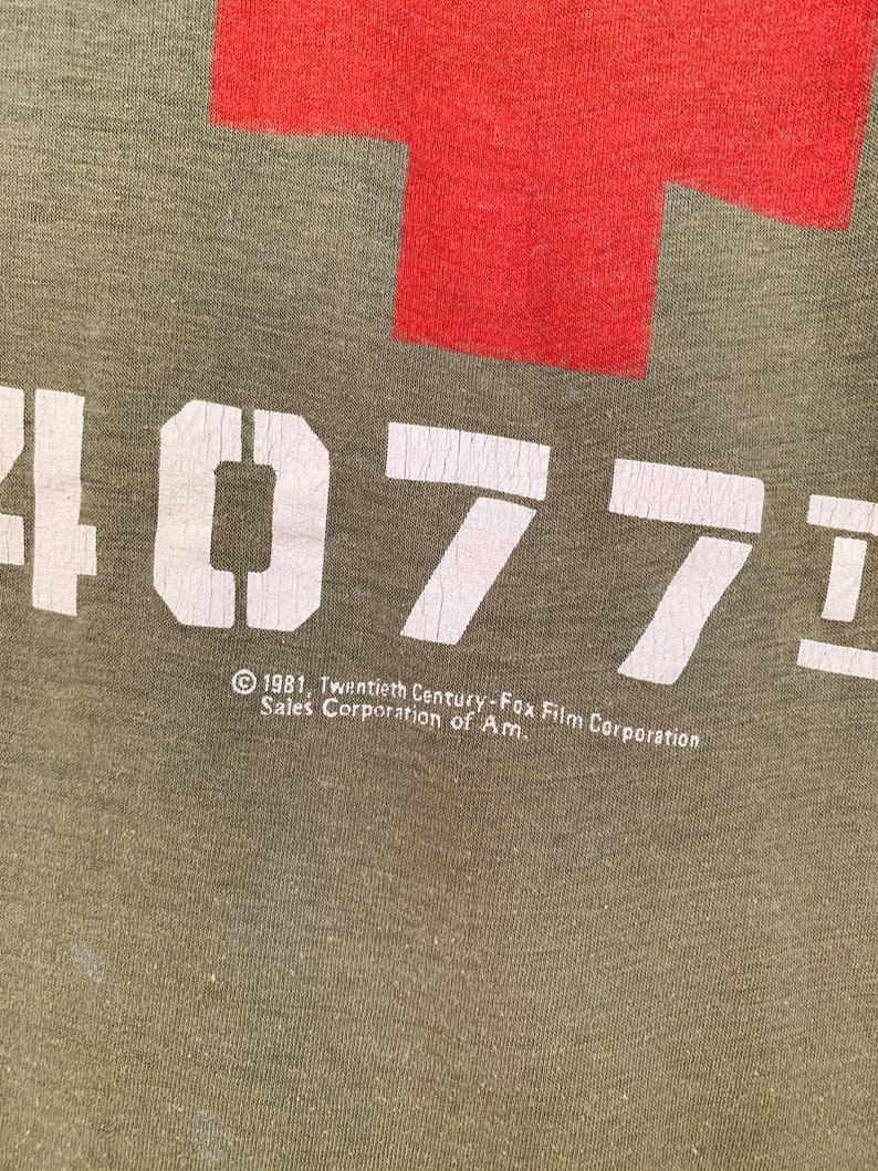 May include: Olive green t-shirt with a large red cross and the white text "4077". Below the text is the copyright information: "&copy; 1981, Twentieth Century-Fox Film Corporation Sales Corporation of Am."