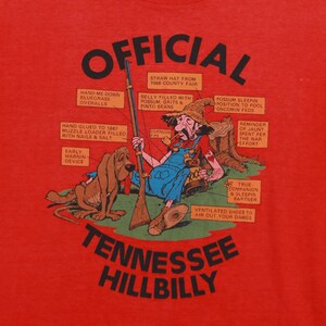 May include: A red t-shirt with a cartoon illustration of a man and a dog. The man is wearing a straw hat, overalls, and a corncob pipe. The dog is a bloodhound. The text "OFFICIAL TENNESSEE HILLBILLY" is printed in black letters on the shirt.