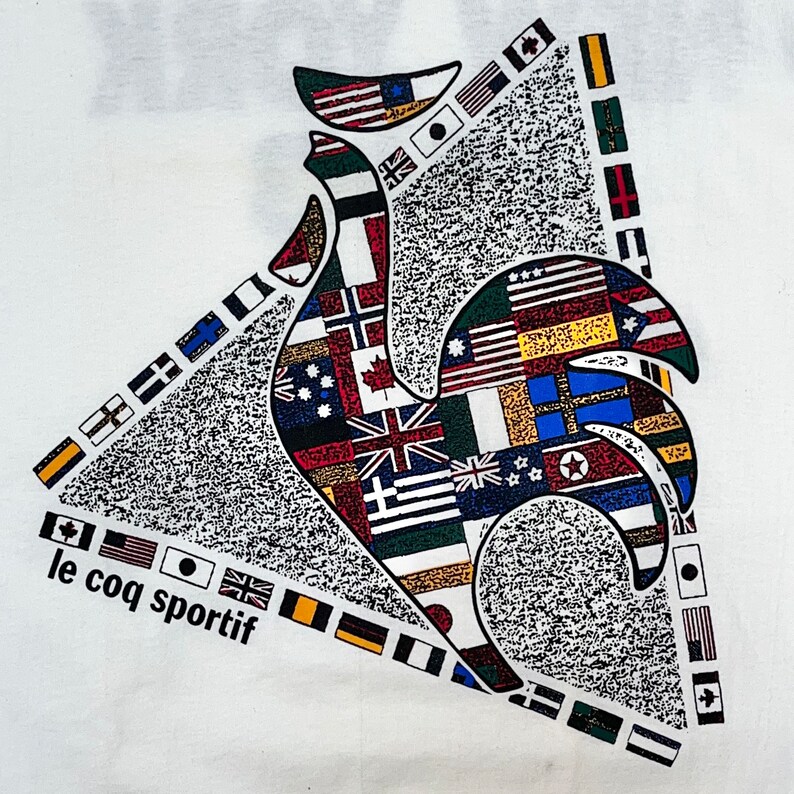 May include: A white t-shirt with a rooster design made up of flags from different countries. The rooster is in the center of the shirt and the text "le coq sportif" is at the bottom left corner.
