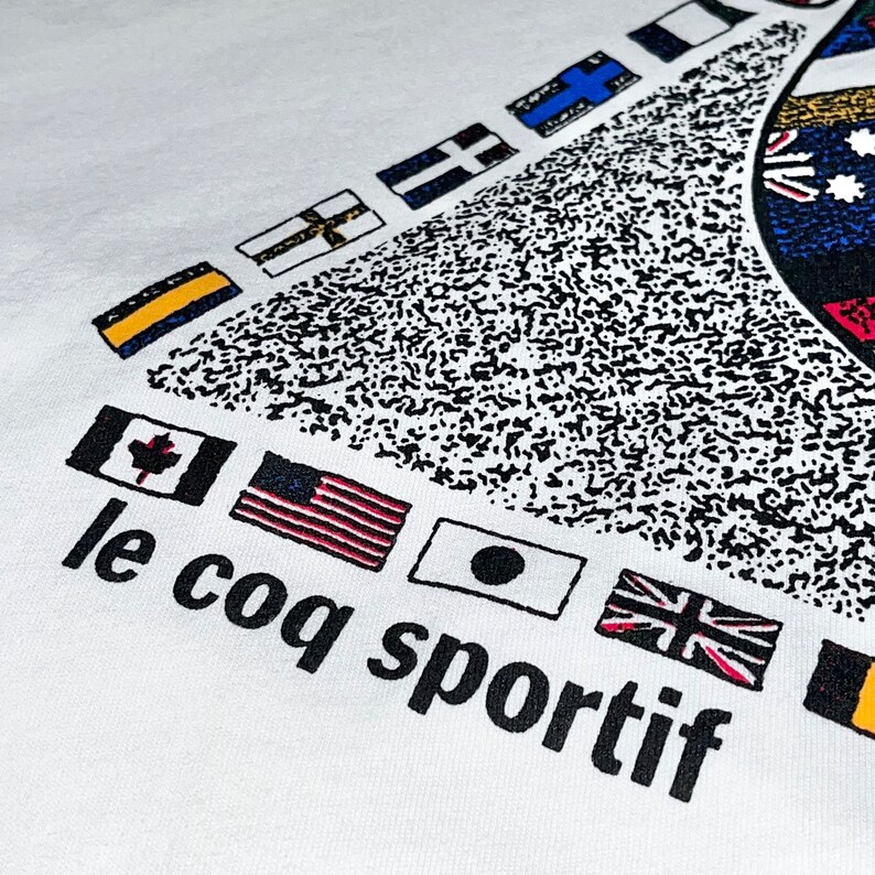 May include: White t-shirt with a graphic of flags from different countries arranged in a triangle shape. The text "le coq sportif" is printed below the flags.