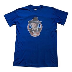 May include: A blue t-shirt with a picture of Johnny Rogers in a cowboy hat and a framed portrait. The text "Johnny Rogers" is visible on the shirt.