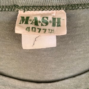 May include: Close-up of a green t-shirt with a tag that reads "MASH 4077TH" in green lettering. The tag is off-white with a small tear. The shirt's fabric is a solid, muted green color.