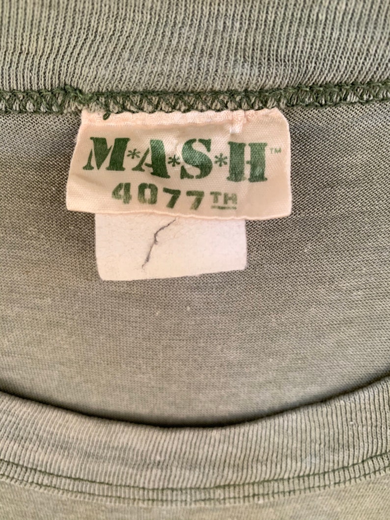 May include: Close-up of a green t-shirt with a tag that reads "MASH 4077TH" in green lettering. The tag is off-white with a small tear. The shirt's fabric is a solid, muted green color.