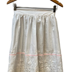 May include: White cotton apron with eyelet lace trim and pink ribbon accents. The apron has a gathered skirt and a simple waistband.