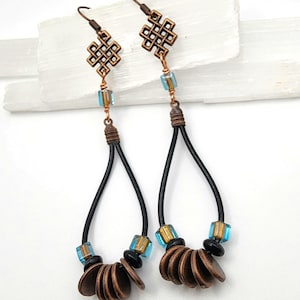 May include: A pair of dangle earrings featuring a copper-toned eternal knot charm, a single blue and gold square bead, and a black leather cord loop. The base of each loop is adorned with small black beads and a cluster of round copper-toned discs.