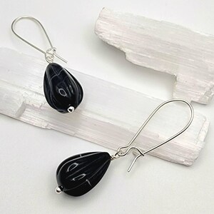 May include: A pair of drop earrings featuring polished black teardrop-shaped beads with vertical fluted detailing. Each bead is suspended from a simple silver-toned metal hook wire, creating a clean and balanced design for these dangling accessories.