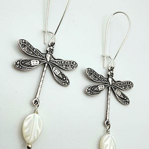 May include: A pair of silver-toned dragonfly earrings featuring intricate filigree patterns on the wings and a long, segmented body. Each earring hangs from a simple silver hook and is finished with a single, white, leaf-shaped mother-of-pearl bead.