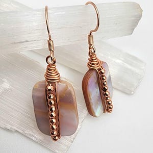 May include: A pair of handcrafted earrings featuring rectangular, iridescent stones. The stones are wrapped in copper wire with a spiral design at the top and a line of small copper beads along the side. The earrings hang from copper ear wires.