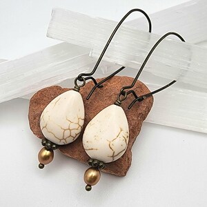 May include: A pair of drop earrings featuring teardrop-shaped white stones with brown veining, accented by small bronze-toned beads and dark metal ear wires. The earrings hang from a pair of curved hooks, showcasing a contrast between the light stone and the metallic components.
