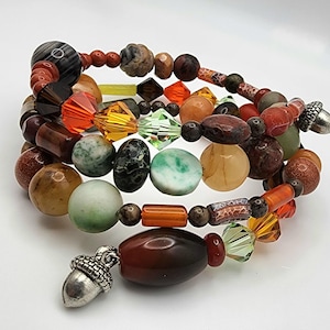 May include: A stack of beaded bracelets in various colors, including brown, orange, green, and yellow. The bracelets feature a variety of bead shapes and sizes, with a silver acorn charm dangling from one. The beads appear to be made of stone and glass.