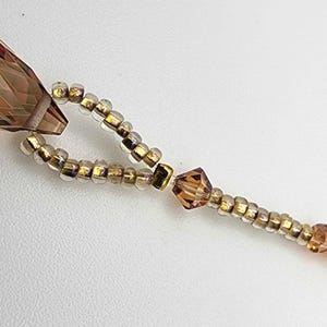 May include: A close-up of a beaded necklace with a teardrop-shaped crystal pendant. The beads are a mix of gold and clear, and the crystal is a deep amber color.