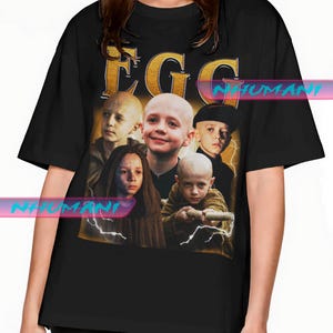 Könnte beinhalten: Schwarzes T-Shirt mit einem Aufdruck von mehreren Kindern mit rasierten Köpfen, dem Wort "EGG" in Goldbuchstaben und Blitz-Akzenten. Das Design hat einen Vintage-Look.