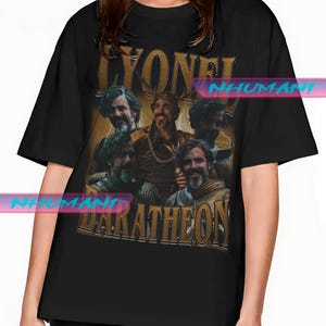 Könnte beinhalten: Schwarzes T-Shirt mit einem Retro-Grafik von mehreren Männern, dem Text "LYONEL" oben und "BARATHEON" unten. Die Grafik ist in Sepia-Tönen mit Blitzakzenten gehalten.