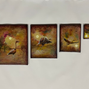 May include: A set of four framed art pieces depicting herons in a wetland scene. The paintings feature a warm color palette with a full moon and a heron in various poses, including standing, taking flight, and landing.