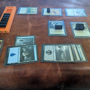 May include: An orange plastic card counter with black dice, and several Magic: The Gathering cards. The cards are in protective sleeves and feature various illustrations and symbols. Some dice show the numbers +21+2 and +8+5.
