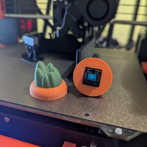 May include: Two 3D-printed items: a green mountain-shaped object on an orange base and an orange circle with a blue square component. The items are on a grey surface, likely a 3D printer bed. The objects are likely components for a mechanical keyboard.