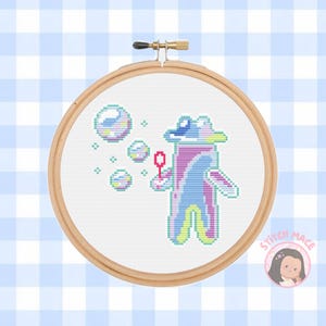 May include: Cross stitch pattern of a purple and blue creature blowing bubbles with a red wand. The creature is wearing a hat and has a pink and blue body. The background is white with a few small blue and green bubbles.