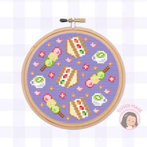 Może przedstawiać: Wzór haftu krzyżykowego w kolorze fioletowym z różnymi japońskimi przekąskami, w tym ciastem, mochi i zieloną herbatą. Wzór jest otoczony drewnianym obręczą do haftu.