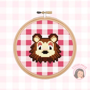 May include: Cross-stitch embroidery hoop with a pink and white gingham pattern. The centre of the hoop features a brown bear with pink cheeks and a brown nose.