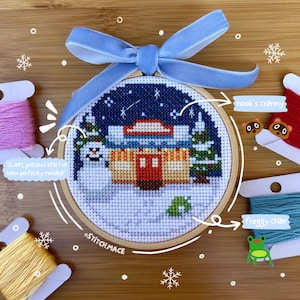 Puede incluir: Un adorno de bordado de aro cosido a mano que presenta una escena invernal con una casa, un muñeco de nieve y un pino. El aro es de madera y tiene una cinta azul atada. El texto "Sweet, precious life! I've been perfectly molded!" y "STITCH.MACE" están escritos en la imagen.