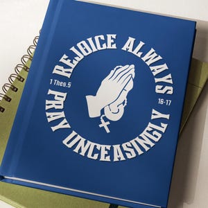 Puede incluir: Un cuaderno azul con las palabras "REJOICE ALWAYS PRAY UNCEASINGLY" que rodean un gráfico de manos rezando. Debajo hay un cuaderno verde. Los cuadernos están sobre una superficie blanca.