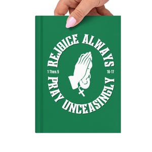 Puede incluir: Un diario verde con las palabras "Alegraos siempre, orad sin cesar" rodeando un gráfico de manos rezando. El texto también incluye "1 Tes. 5:16-17". El diario se sostiene en una mano.