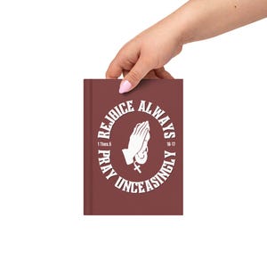 Puede incluir: Un diario de color granate con las palabras "Rejoice Always, Pray Unceasingly" que rodean un gráfico de manos rezando. El texto también incluye "1 Thes. 5" y "16-17" en blanco. El diario es sostenido por una mano.