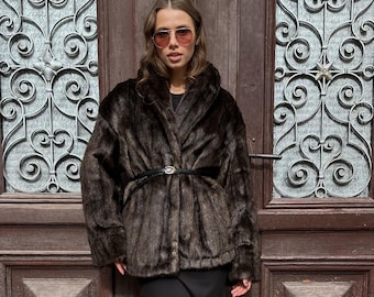 May include: A woman wearing a brown fur coat with a black belt, black pants, and leopard print sneakers. She is standing in front of a wooden door with a decorative metal grate.