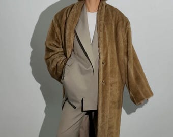 Op de afbeelding: Een persoon draagt een bruine imitatiebontjas over een lichtbruin pak met een wit overhemd. De persoon draagt ook een zwarte zonnebril. De jas is lang en heeft knopen. Het pak bestaat uit een blazer en een broek met wijde pijpen.