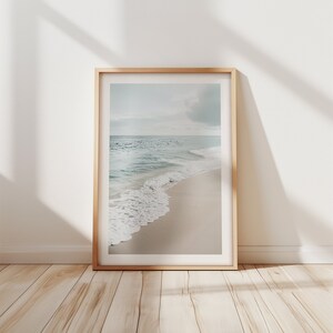 May include: A framed print showing a tranquil beach scene. The image captures a sandy shore with soft waves from the turquoise sea under a cloudy sky. The frame is made of light-coloured wood.