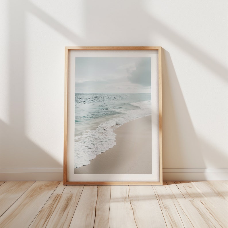 May include: A framed print showing a tranquil beach scene. The image captures a sandy shore with soft waves from the turquoise sea under a cloudy sky. The frame is made of light-coloured wood.