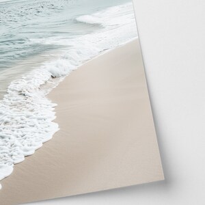 May include: A tranquil beach vista featuring pale beige sand and calm waves. The sea shifts from a light turquoise to a deeper blue, with white foam from the breaking waves. The sky is overcast.