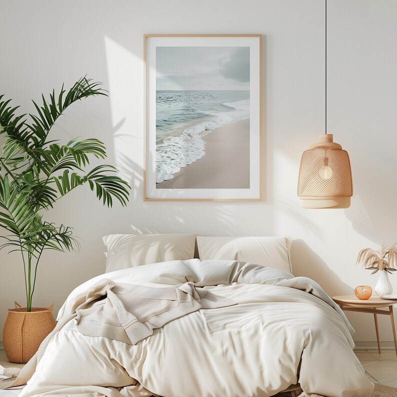 May include: A framed print of a beach scene hangs above a bed with cream bedding. A woven pendant light hangs from the ceiling. A potted palm tree sits in a woven basket. The room has a neutral, calming aesthetic.