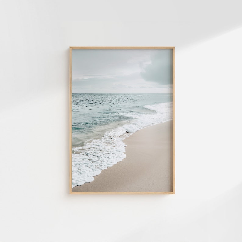 May include: A framed print depicting a beach scene. The image shows a sandy beach with waves gently breaking on the shore. The water is a pale blue-green, and the sky is cloudy. The frame is a light wood colour.