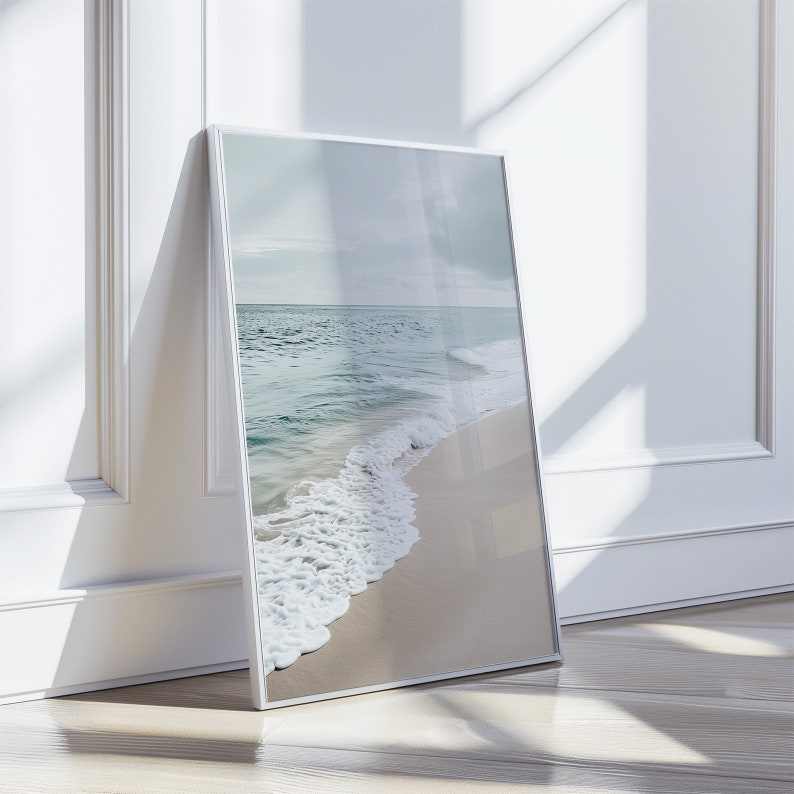 May include: A framed print depicting a beach scene. The image shows a sandy shore with waves breaking on the coast from the turquoise sea. The sky is overcast, and the print is in a silver frame, leaning against a white wall.