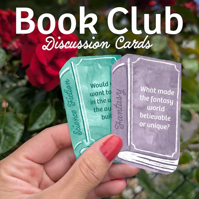 May include: A hand holds two discussion cards shaped like books. The cards are labeled "Science Fiction" and "Fantasy" with discussion prompts. The background includes the text "Book Club Discussion Cards" and a blurred floral arrangement.