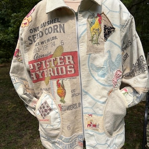 May include: A beige zip-up jacket constructed from repurposed feed sacks featuring printed text including ONE BUSHEL SEED CORN and PFISTER HYBRIDS. The garment displays rooster illustrations, a chicken wire pattern, and two front patch pockets.