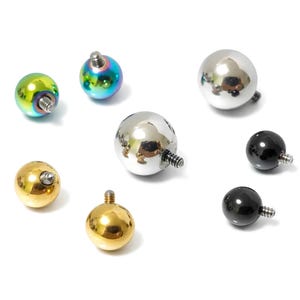 Op de afbeelding: Een assortiment bolvormige sieraden voor piercings in goud, zilver, zwart en iriserende kleuren. Elke bal heeft een schroefdraad voor bevestiging. De sieraden worden gepresenteerd op een witte achtergrond.