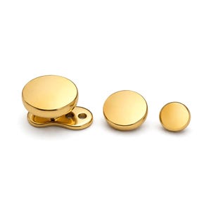 Può includere: Una collezione di gioielli rotondi color oro. Il set comprende tre pezzi: due borchie circolari e un pezzo più grande con una base curva unica con due fori. I gioielli hanno una superficie lucida e riflettente.