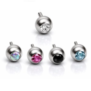 Op de afbeelding: Een verzameling van zes zilverkleurige, draadbolvormige lichaamsjuwelen. Elk stuk heeft een verschillend gekleurde edelsteen: helder, iriserend, roze, zwart en lichtblauw. De sieraden zijn ontworpen voor piercings.