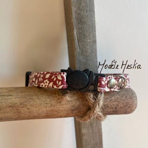 Peut inclure: Un collier pour chat à motif floral rouge et blanc avec une boucle noire en forme de chat et une boucle de sécurité argentée. Le collier est sur une surface en bois. "Modèle Heshia" est écrit en blanc sur fond noir.
