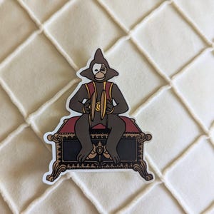 Può includere: Un adesivo raffigurante un personaggio di scimmia dei cartoni animati con una mezza maschera, che tiene dei piatti e seduto su un forziere decorativo. La scimmia è marrone con accenti rossi e dorati. Il forziere è nero con finiture dorate e cuscini rossi.