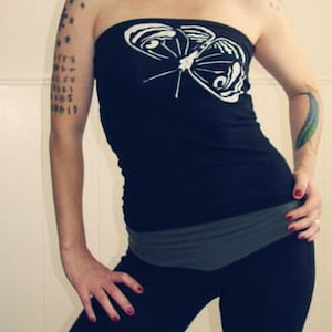 May include: A woman wearing a black tank top with a white butterfly graphic. The tank top has a gray waistband and the woman is wearing black pants.